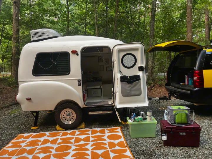 White HC1 rv rental camper & yellow SUV ready for adventure at a forest campsite. An orange mat & camping gear complete the setup.. Happier Camper HC1 2022