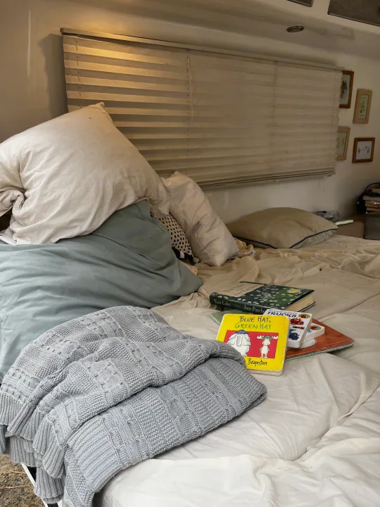 Cozy rv rental bed with white and light blue pillows, grey knitted blanket, a stack of books, and horizontal window blinds.. Happier Camper HC1 2022