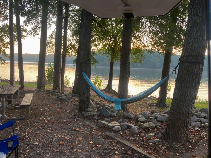 RV rental campsite by a lake, featuring a blue hammock, picnic table, and golden sunrise on the water.. Happier Camper HC1 2022