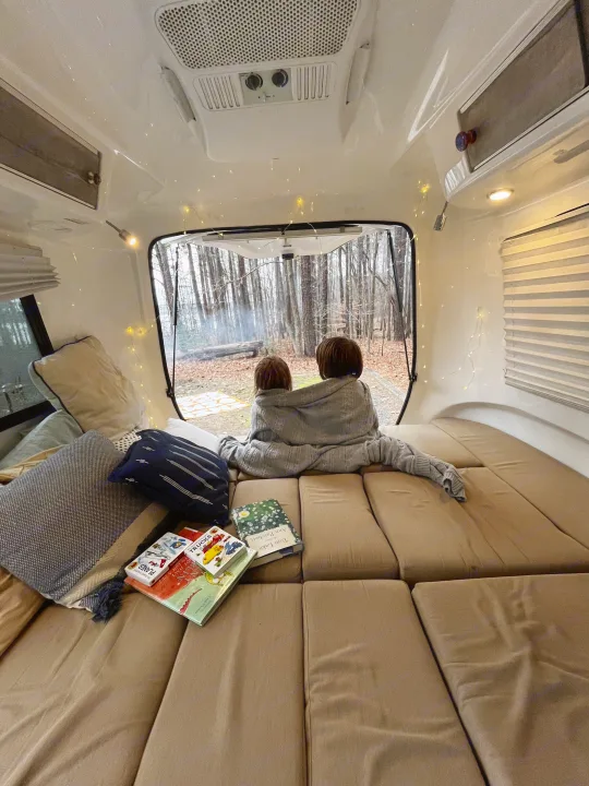 Inside a cozy **rv rental**: two children under a blanket watch a forest campfire. Books and string lights add to the warm atmosphere.. Happier Camper HC1 2022