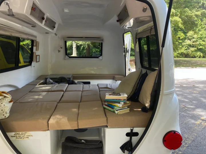 Inside a compact RV rental: a plush bed, stack of books, and smart storage solutions for travelers.. Happier Camper HC1 2022