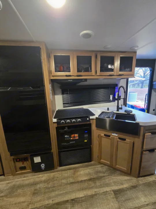 This modern RV rental kitchen boasts black appliances (fridge, microwave, stovetop, farmhouse sink), light wood cabinets, and blinds.. Forest River Cherokee Grey Wolf 2024