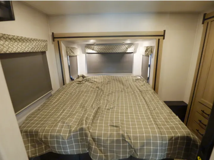 Cozy RV rental bedroom features a plaid bed, overhead storage, window blinds, reading lights, and a wooden wardrobe.. Classe C 24-26 pieds avec extension(s) 2027