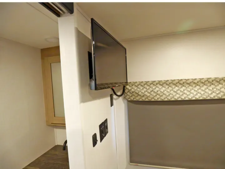 Modern RV rental interior: wall-mounted TV, control switches, and a window with a patterned valance and roller blind.. Classe C 24-26 pieds avec extension(s) 2027