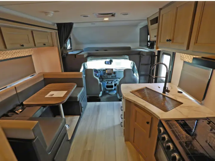 Modern rv rental interior: kitchen with sink/stove, dinette, and bright cockpit. Overhead bunk and wood-look flooring.. Classe C 24-26 pieds avec extension(s) 2027