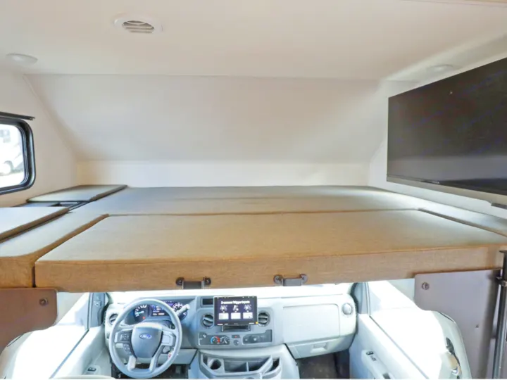 RV rental's cab-over bunk with mattresses and TV, overlooking the Ford dashboard and cockpit area for driving.. Classe C 24-26 pieds avec extension(s) 2027