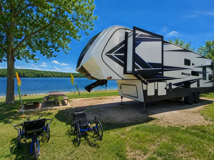 Experience lakefront tranquility with this spacious fifth-wheel RV rental, picnic spot, and recumbent trikes on a sunny day.. Grand Design Momentum 2019