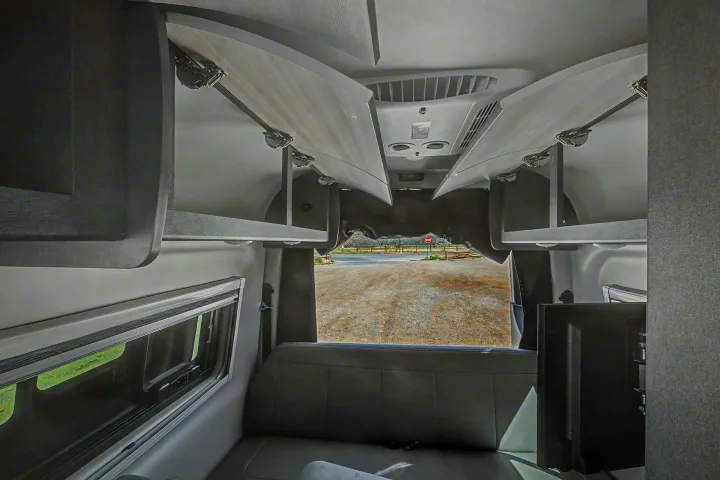 Spacious RV rental interior featuring open overhead storage, bench seating, a window, and a TV, with a view of a gravel lot.. Thor Tranquility 2025