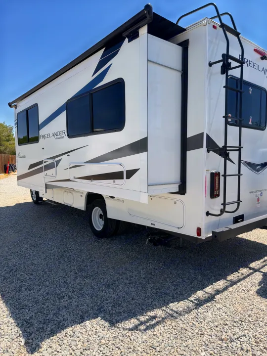 This white Coachmen Freelander RV rental, with its side extended and roof ladder, is parked on a gravel lot under a blue sky.. Forest River Coachmen Freelander 2023