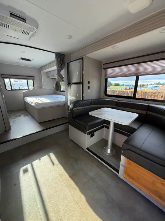 This rv rental's bright interior features an elevated bed, U-shaped dinette, fridge, and windows revealing outdoor views.. Forest River Coachmen Freelander 2023