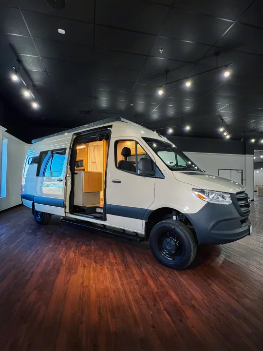 Ready for adventure! This modern beige camper van with an open side door reveals an interior shower, ideal for your RV rental journey.. Mercedes-Benz Sprinter 2024