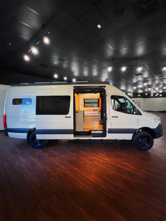 This modern white camper van, with its side door open to a cozy wooden interior, is ready for your RV rental adventure.. Mercedes-Benz Sprinter 2024