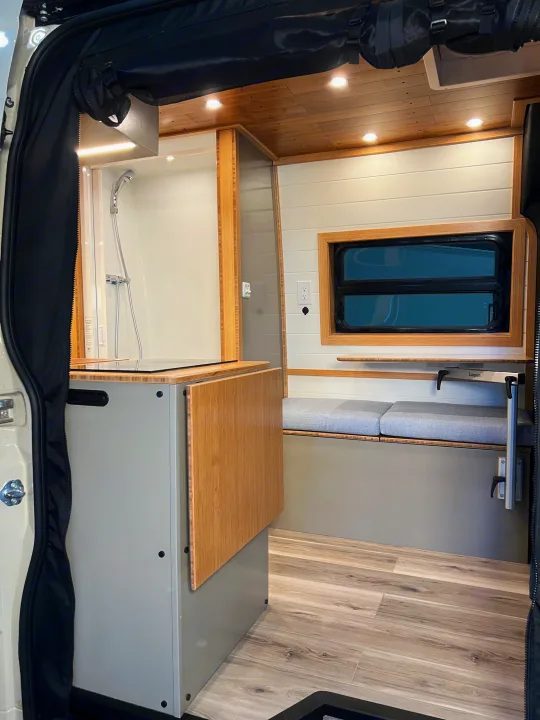 An RV rental interior showing a shower, foldable table, cushioned bench, and window with wood paneling & flooring.. Mercedes-Benz Sprinter 2024