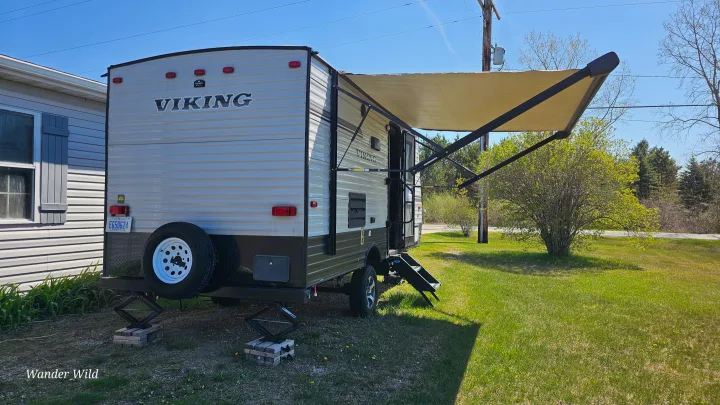 RV rental: A beige travel trailer with an extended awning, stability jacks, and a spare tire set up on a grassy site.. Forest River Viking 2018