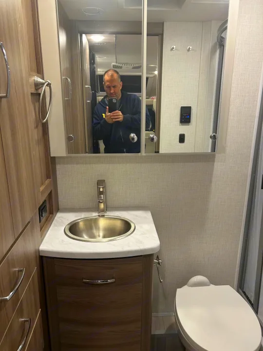 Inside a compact RV rental bathroom: sink, mirrored cabinet, toilet, and shower stall. A person is reflected in the mirror.. Thor Delano 2026