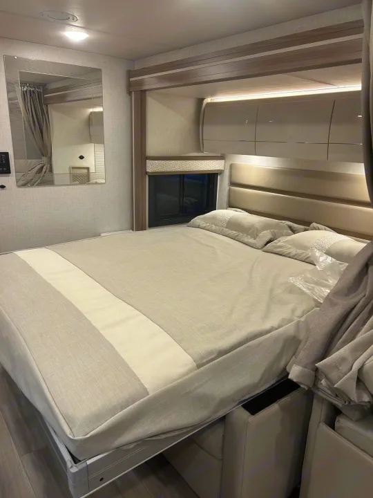 Inviting RV rental bedroom: light-toned bed with padded headboard, sleek overhead cabinets & storage. Bed base is lifted.. Thor Delano 2026