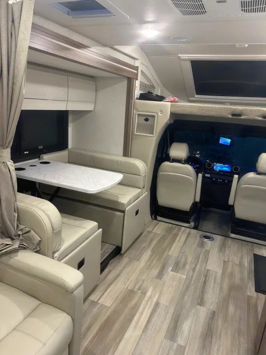 Inside this modern RV rental, observe the light faux leather seating, dining table, wood-look flooring, and front cockpit area.. Thor Delano 2026