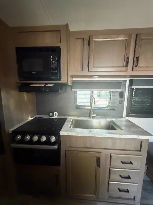 Compact RV rental kitchen featuring a black microwave, gas cooktop, stainless steel sink, and light wood cabinets with counter space.. Starcraft Autumn Ridge 2022