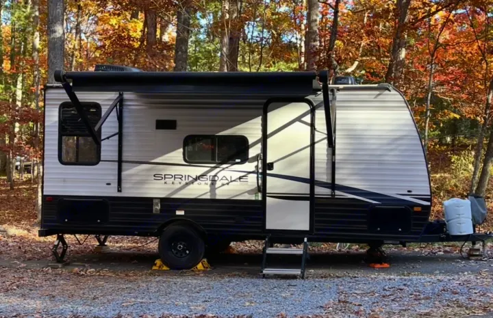 RV rental travel trailer parked at a fall campsite. Its awning is extended, with colorful autumn trees in the background.. Keystone RV Springdale 2023