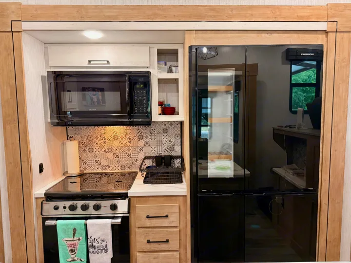 Modern RV rental kitchen featuring a compact setup with microwave, oven, black fridge, and patterned backsplash.. Heartland RVs Milestone 2023