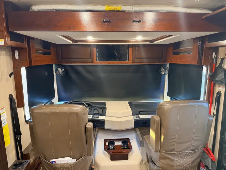 RV rental cockpit view, showing the driver/passenger seats, dashboard, overhead bunk, and windshield with blackout shades.. Fleetwood Bounder 2018
