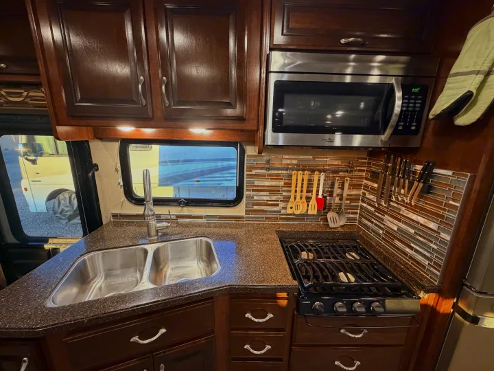 Spacious RV rental kitchen with dark wood cabinets, stainless appliances, double sink, and mosaic backsplash, ready for your travels.. Fleetwood Bounder 2018