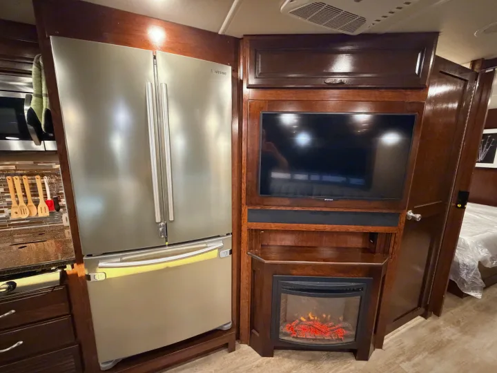 RV rental interior with stainless fridge, TV, & electric fireplace in dark wood cabinetry, and a peek at the bedroom.. Fleetwood Bounder 2018