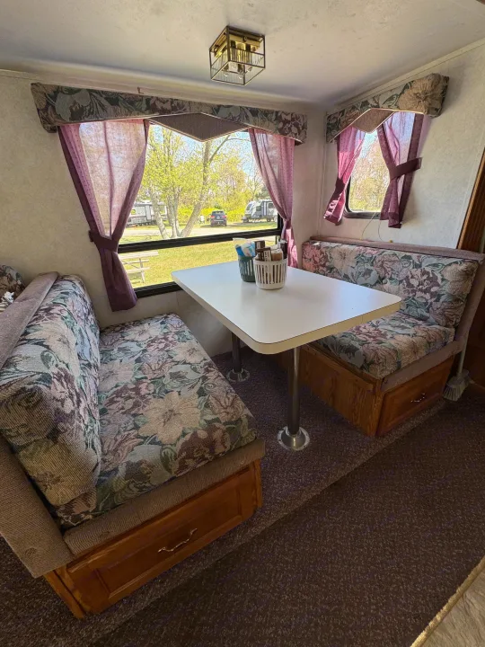 Cozy RV rental dinette with floral cushions, a white table, under-seat storage, and a scenic window view of a campground.. Forest River Wildwood 2000