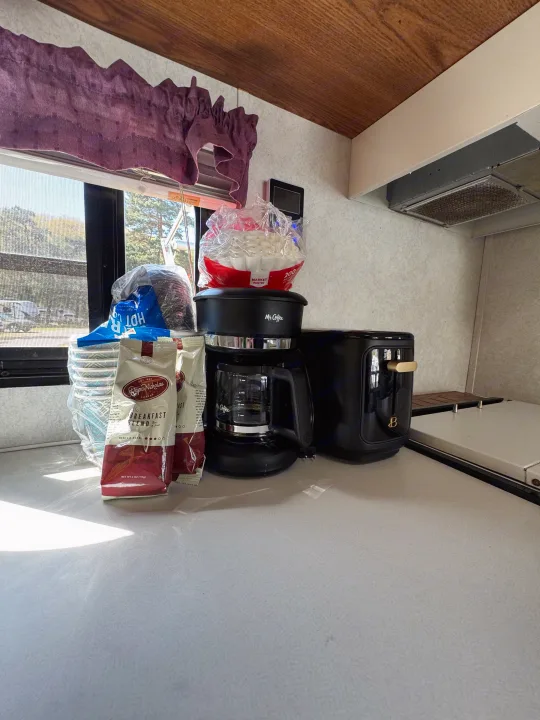 RV rental kitchen counter with a black coffee maker, toaster, coffee bags, filters, and stacked cups. Sunlight streams in.. Forest River Wildwood 2000
