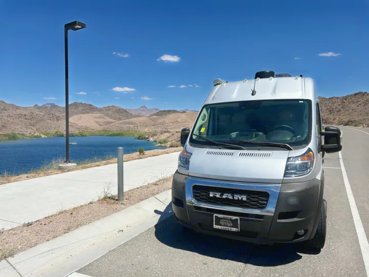 Silver RV rental van parked by a scenic lake with mountains, under a clear blue sky. Ideal for outdoor adventures.. Thor Sequnce 2023