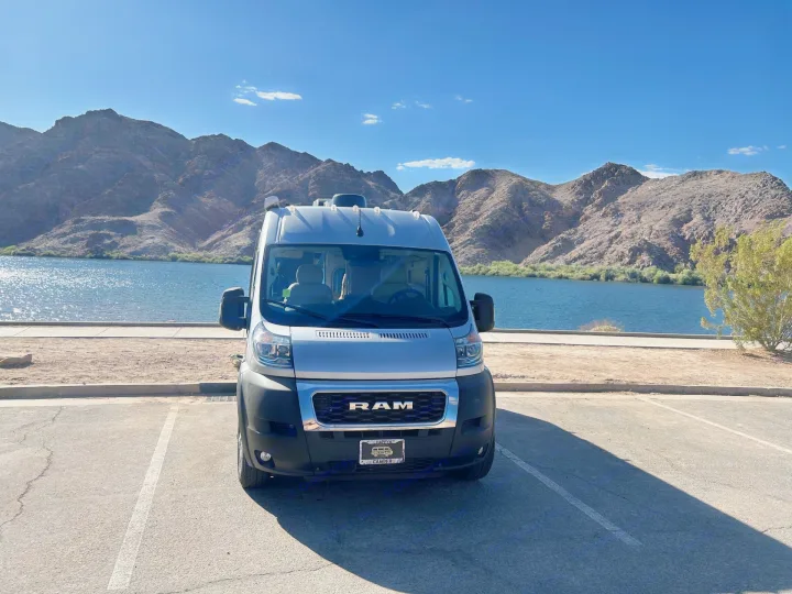 Enjoy your RV rental: a white camper van parked lakeside with majestic mountains under a blue sky.. Thor Sequnce 2023