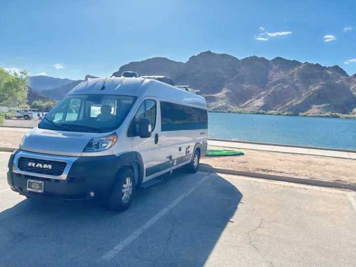 Silver rv rental van parked next to a blue lake and rugged mountains, under a bright sky. Perfect for outdoor travel.. Thor Sequnce 2023