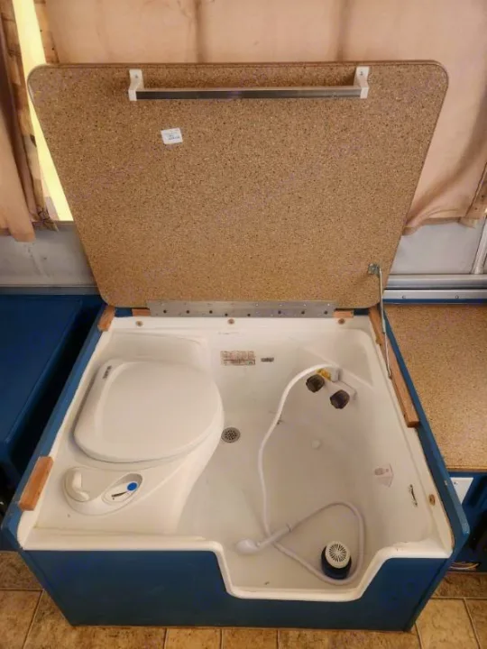 RV rental's compact bathroom: toilet, shower, and propped-open speckled lid with towel bar revealed.. Forest River Rockwood Freedom 2009