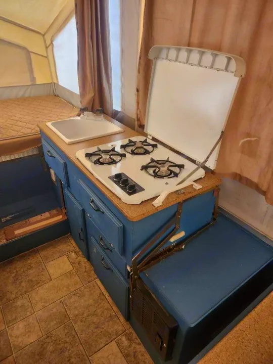 This RV rental features a compact pop-up camper kitchen with a sink, open gas stove, and blue storage cabinets.. Forest River Rockwood Freedom 2009