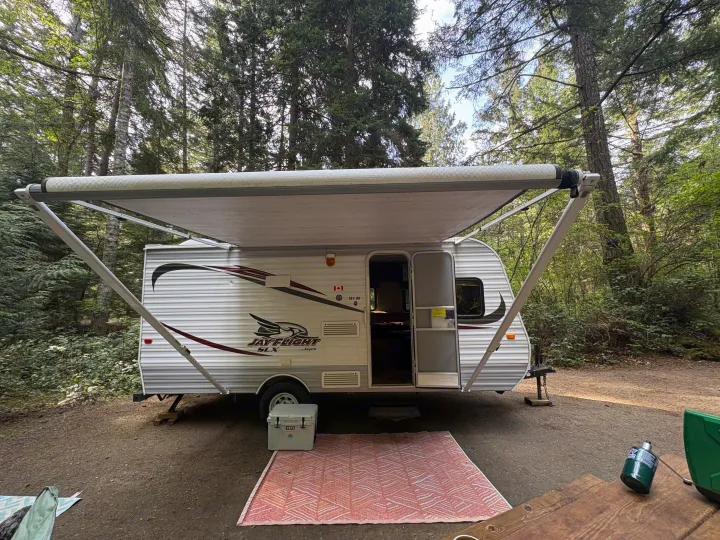 A white Jayco travel trailer, often used as an RV rental, is set up with its awning extended at a shaded forest campsite. Ready for camping!. Jayco Jay Flight 2015