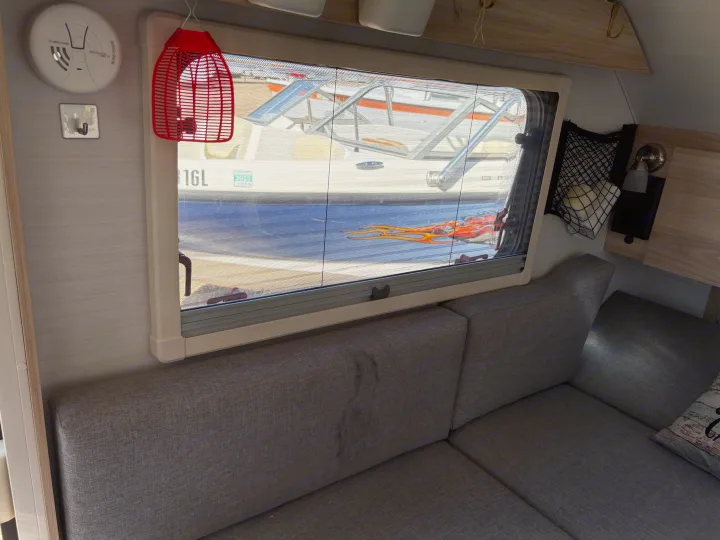 Inside an RV rental: a gray couch, a window offering views of a boat/RV with a solar panel, and a smoke detector.. nuCamp T@B 2024