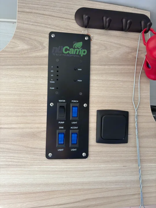 nuCamp RV rental control panel shows tank levels, water pump, and lighting switches. A coat rack is visible above.. nuCamp T@B 2024