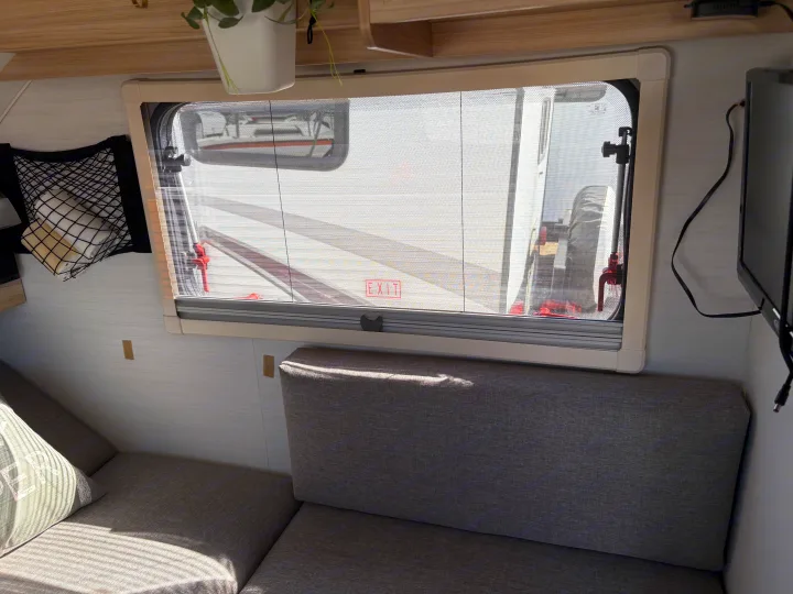 Cozy RV rental interior: bright window with screen, soft gray seating, storage net, and TV. Enjoy comfort on your road trip.. nuCamp T@B 2024