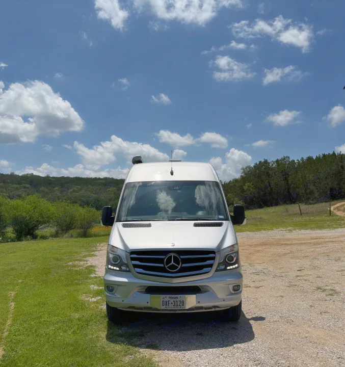 Silver Mercedes-Benz Sprinter van, an RV rental, parked on gravel with a nature backdrop under a cloudy blue sky.. Airstream Interstate 2014