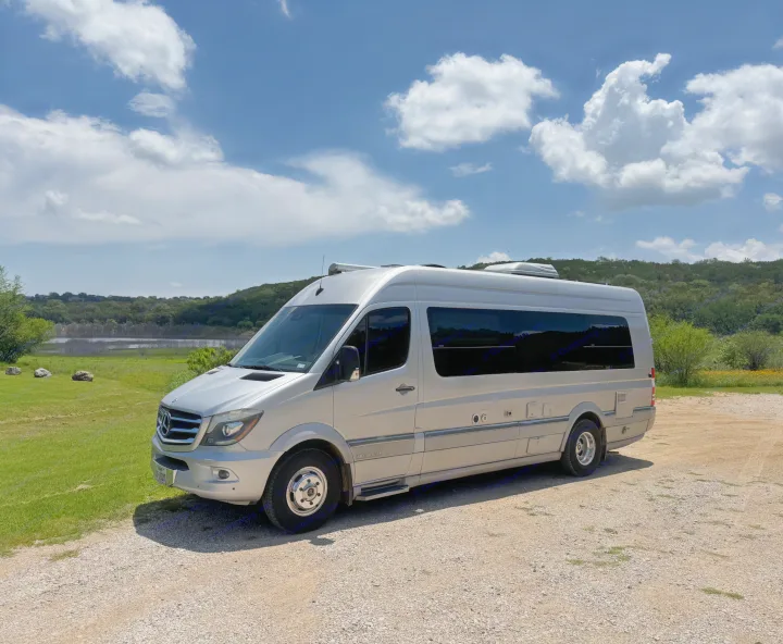 A silver Mercedes Sprinter RV rental, a sleek camper van, parked on gravel with a lake, trees, and cloudy blue sky in background.. Airstream Interstate 2014