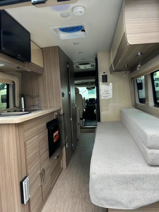 RV rental interior: Modern camper van layout with light wood cabinetry, kitchenette, mounted TV, and a comfy light grey sofa.. Jayco Swift Li 2023