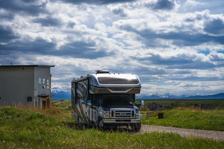 This RV rental, a Jayco motorhome, sits on a gravel path. A modern house, green hills, and snow-capped mountains frame the scenic view.. Jayco Greyhawk Prestige 2021