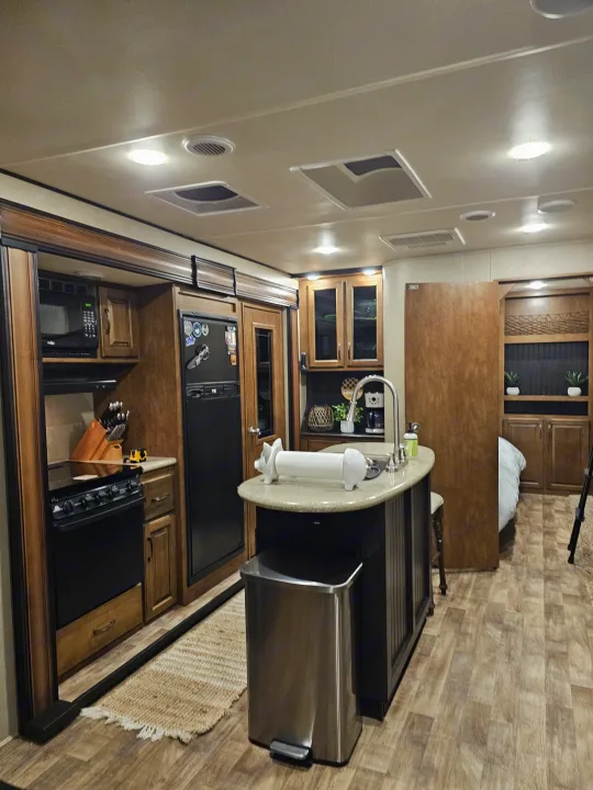 Inside a cozy RV rental: kitchen with stove, fridge, island, and glimpse of the bedroom. Wood floors and warm lighting.. Grand Design Reflection 2016