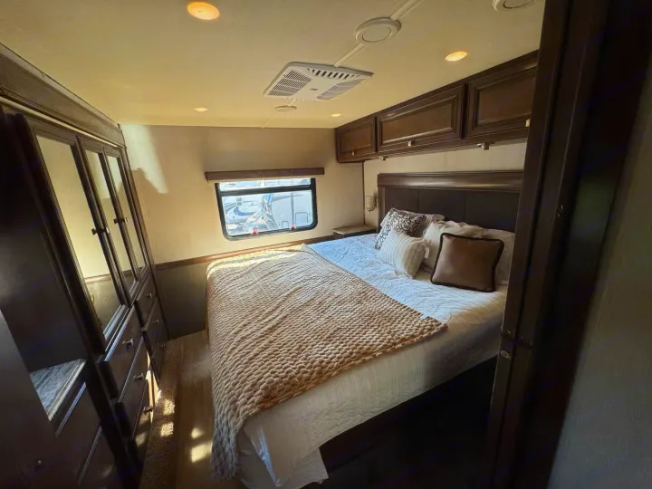 Cozy RV rental bedroom features a comfortable bed, overhead cabinets, and a window for scenic views, ensuring a relaxing travel experience.. Holiday Rambler Admiral 2019
