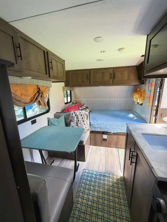 RV rental interior: a cozy bedroom with bed, a dining booth and table, plus a compact kitchenette. Features wood cabinets & light floor.. Coachmen 17BH 2022
