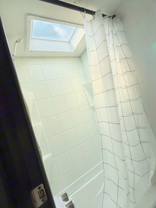 Inside an RV rental shower, featuring a bright skylight, white tiled walls, and a modern black grid shower curtain.. Coachmen 17BH 2022