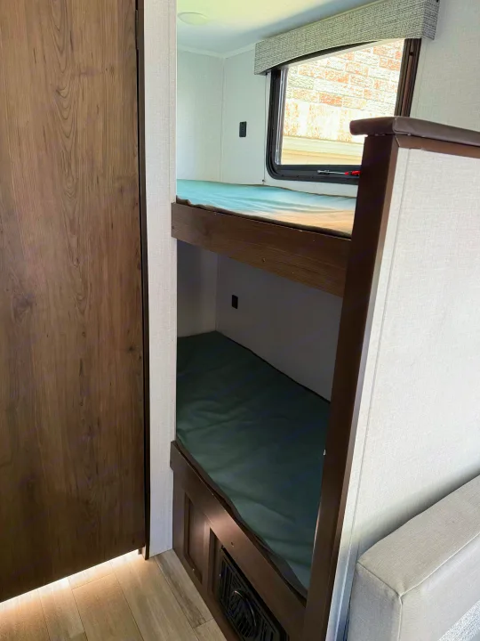 RV rental interior: two bunk beds with dark green mattresses, a window revealing a brick wall, and a wood-textured door.. Coachmen 17BH 2022
