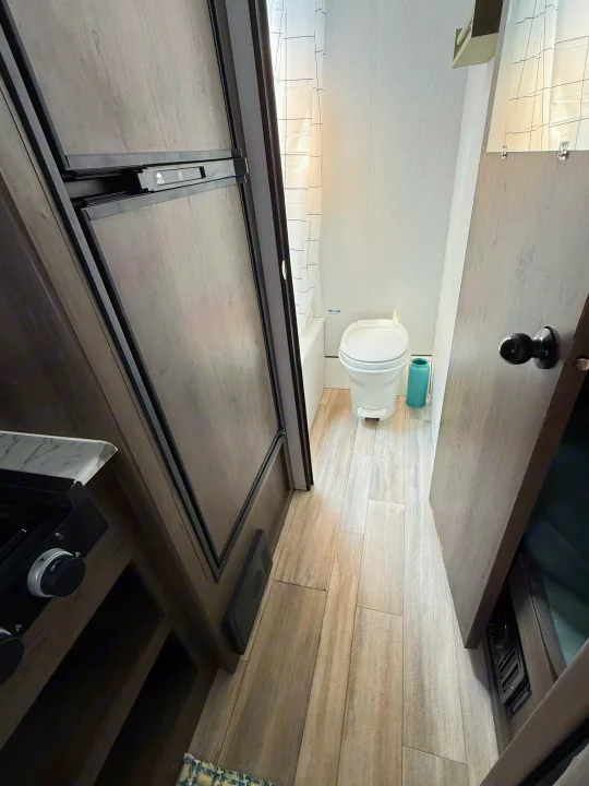 This rv rental interior features a compact bathroom with a white toilet and shower, next to a kitchen with a fridge and stove.. Coachmen 17BH 2022