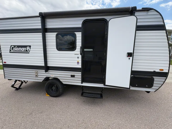 This Coleman travel trailer RV rental, with its door open, awning rolled, and stabilization jacks deployed on asphalt, awaits adventure.. Keystone RV Coleman 2025