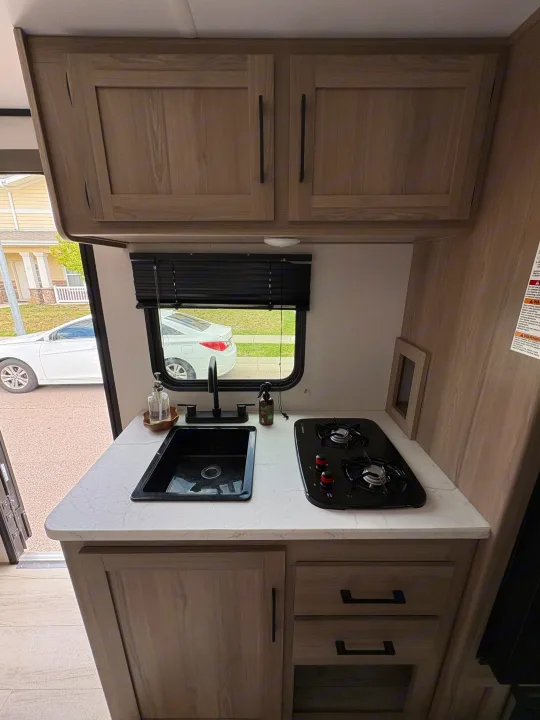 Modern RV rental kitchen with a black sink, two-burner stove, light wood cabinets, and clean white countertops.. Keystone RV Coleman 2025